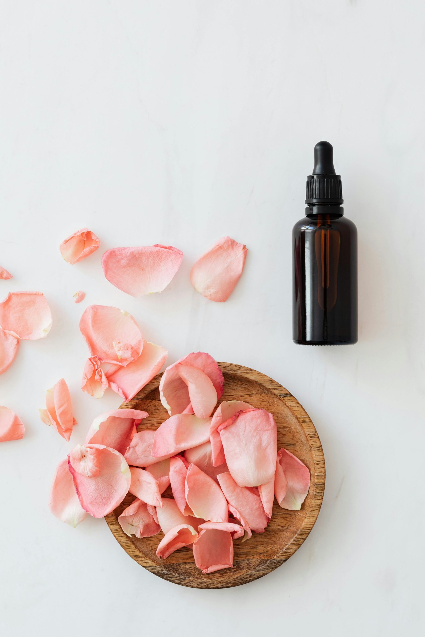 Flat lay of pink rose petals and dark glass essential oil bottle on white background.